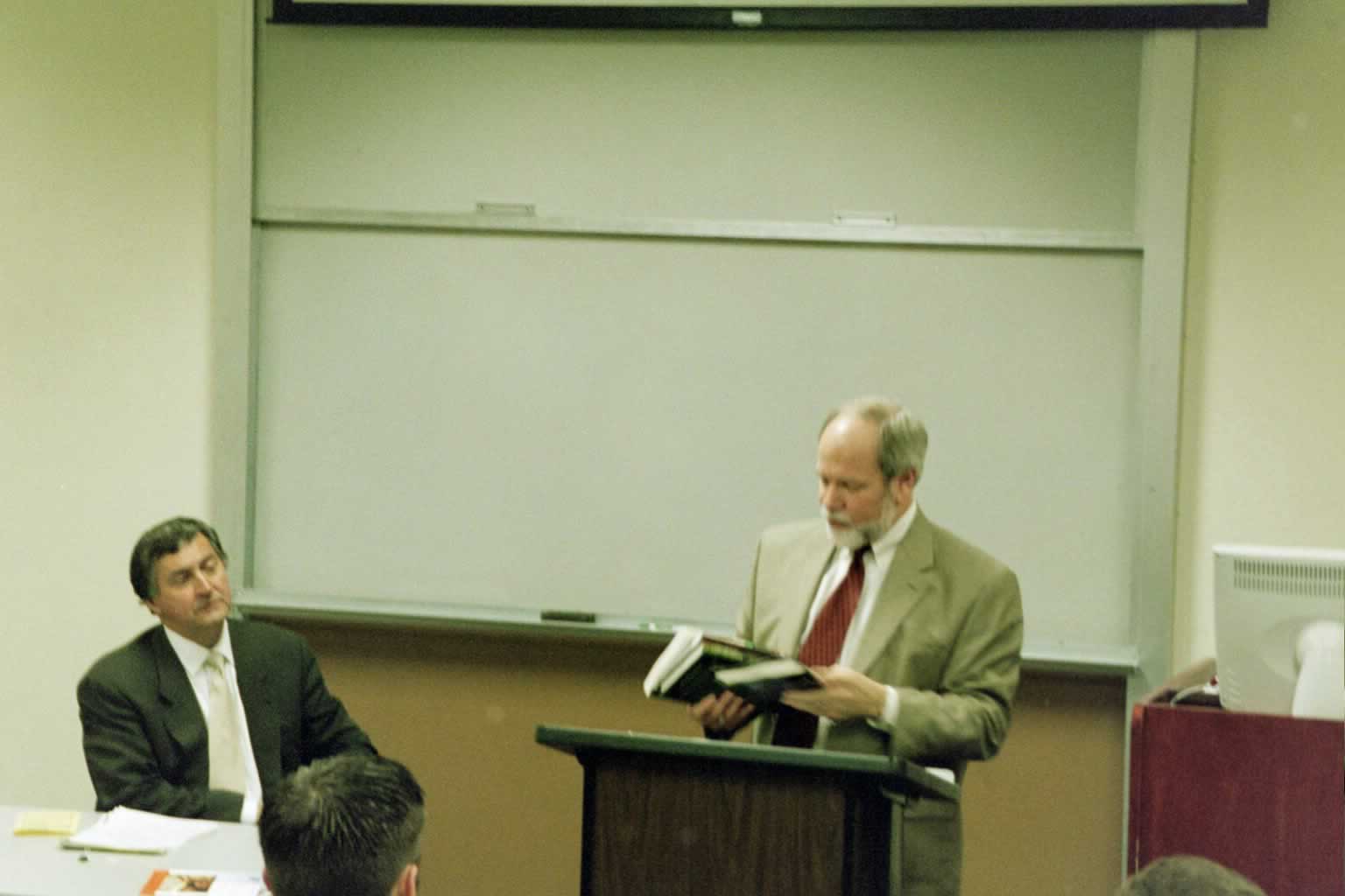 Dr. Naugle standing in front of the podium speaking with Paul Marshall sitting next to him