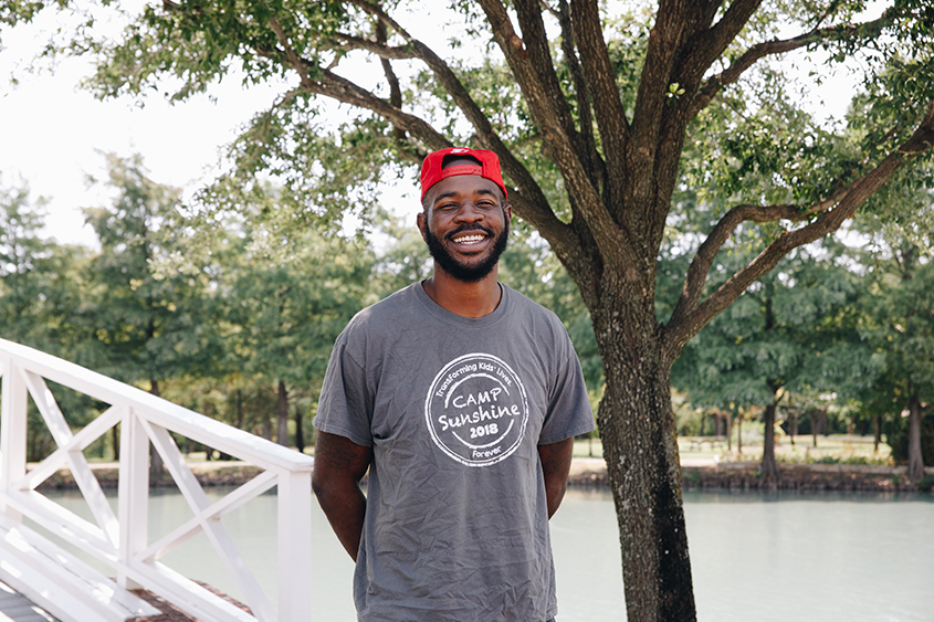 Derrick smiles as he poses in front of Bush Pond with a camp sunshine t-shirt on