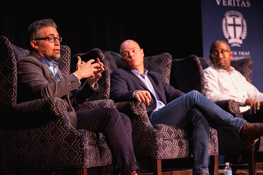 Three men are sitting in chairs on stage. 