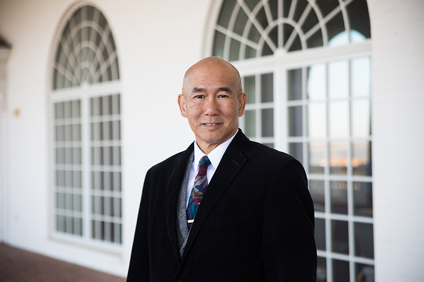 Russel Chun is standing in front of a set of windows smiling. 