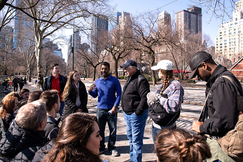 A group of students listen to a lecture in the middle of New York City. 