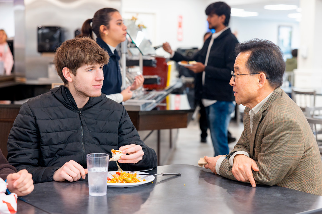 College student eating with a professor in a university dining hall with people in the background