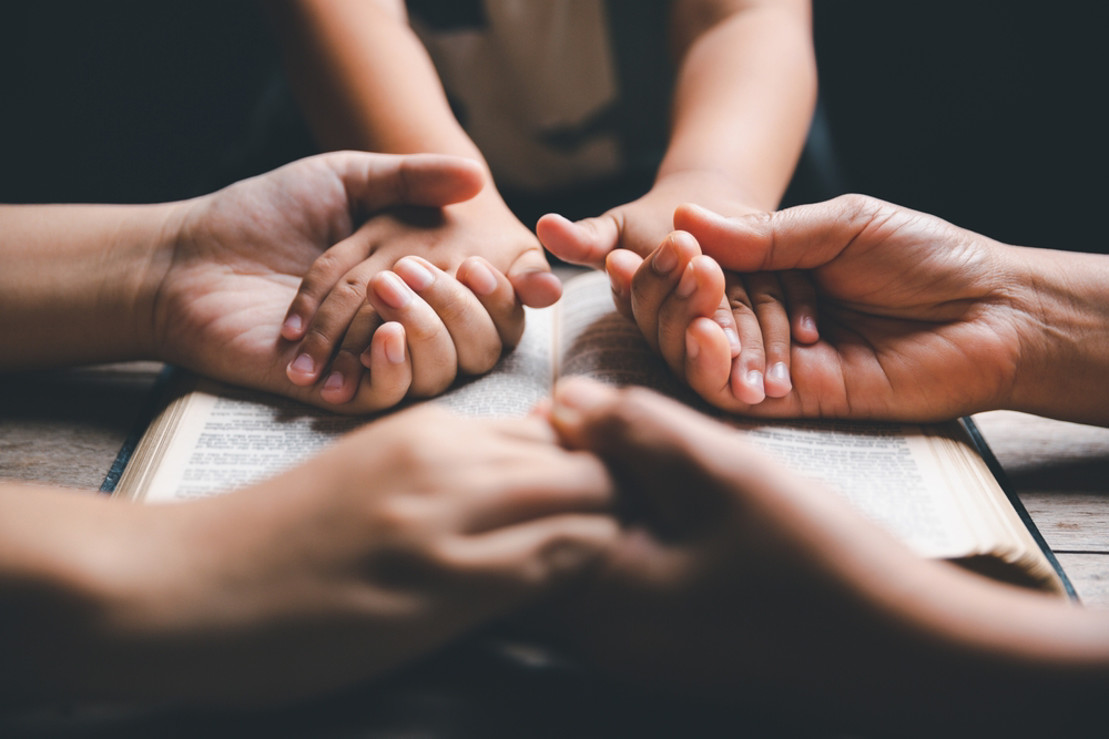 two adults holding hands with a small child praying over an open Bible