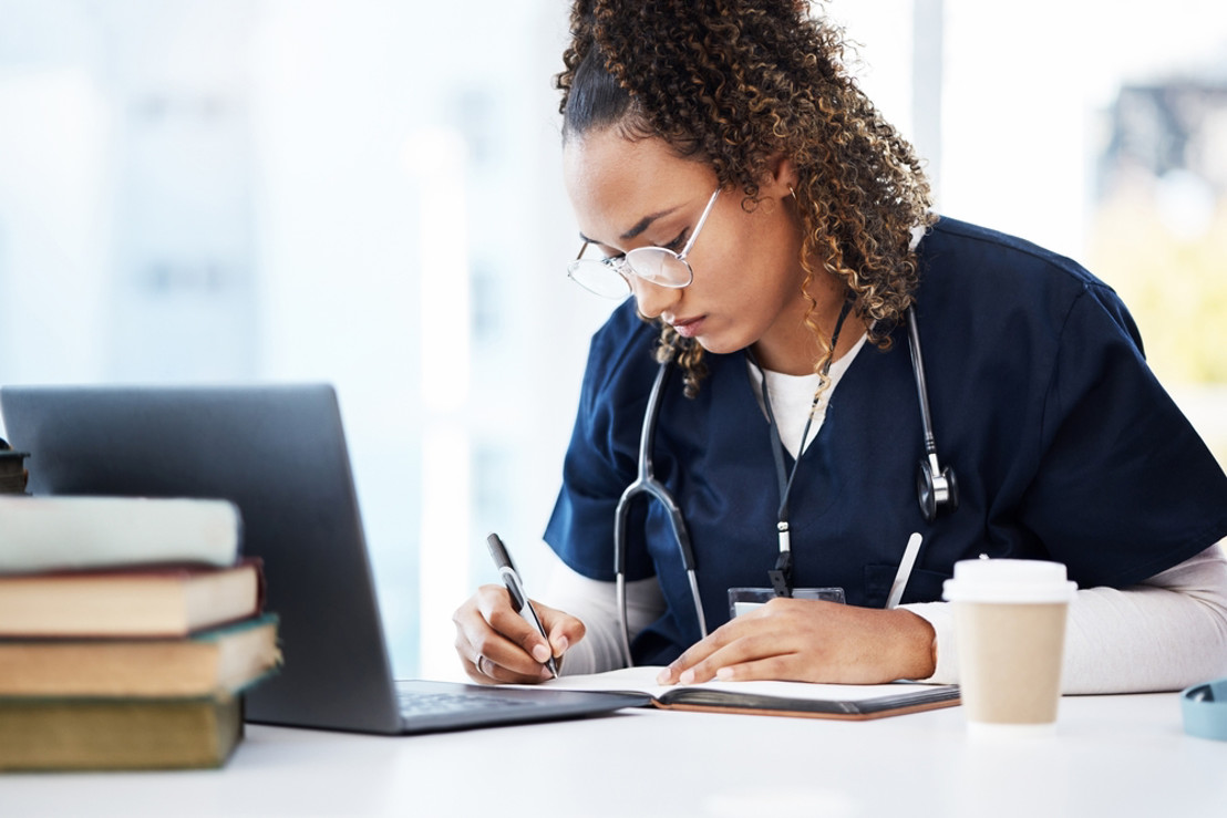 woman nurse writing notes at work while wearing navy scrubs with coffee, laptop, and books on table