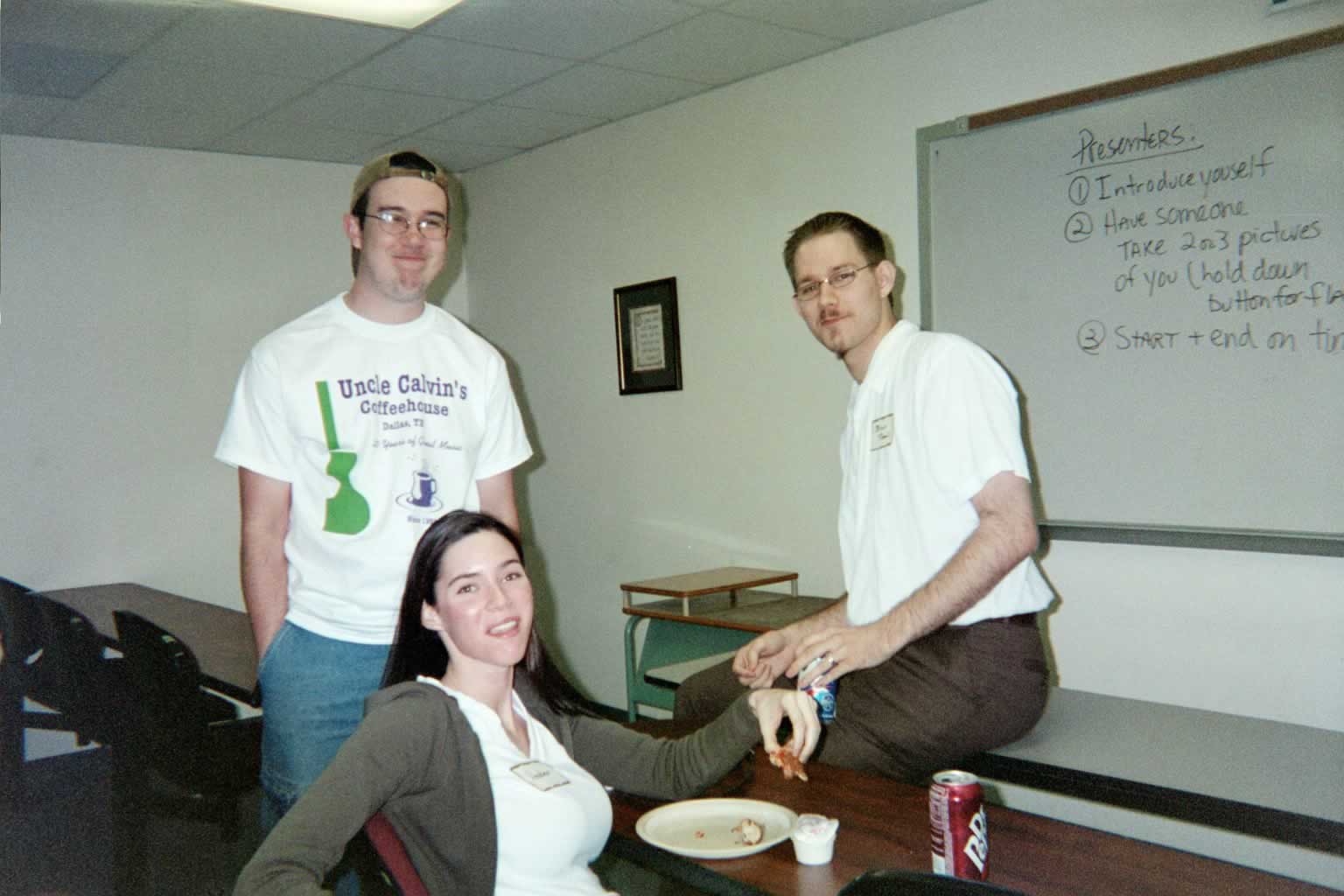picture of two men and a woman standing and sitting while eating