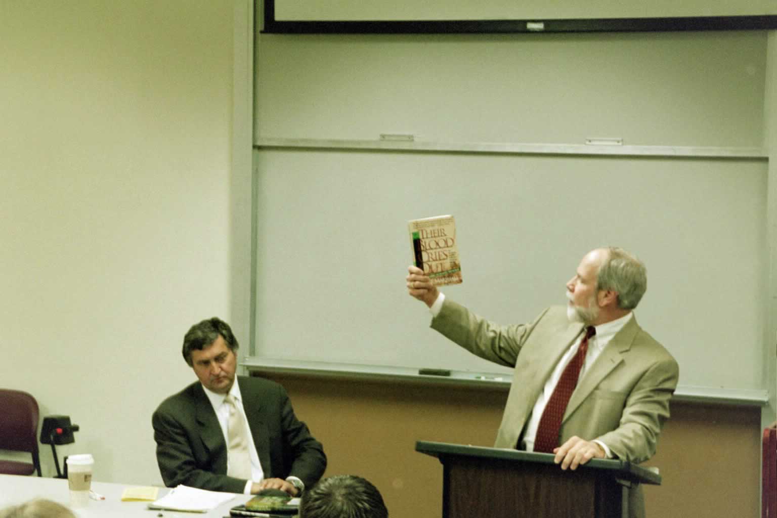 Dr. Naugle standing behind a podium holding up a book with Paul Marshall sitting next to him