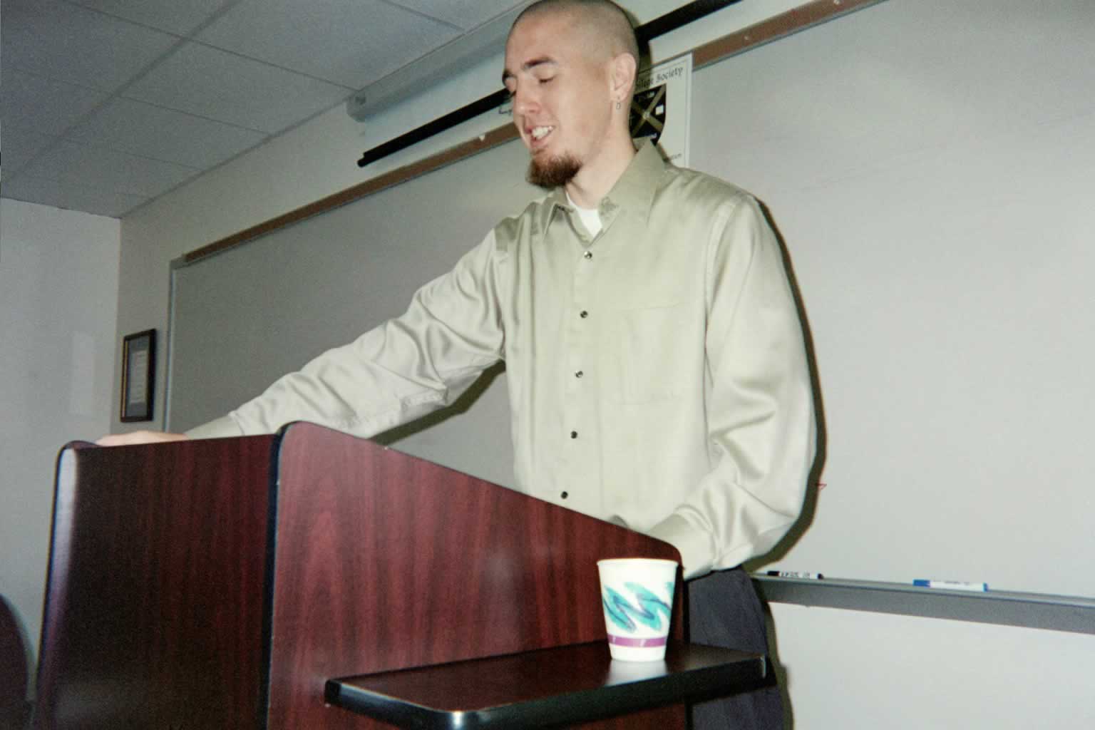 picture of a man standing behind a podium with a cup on the table next to him
