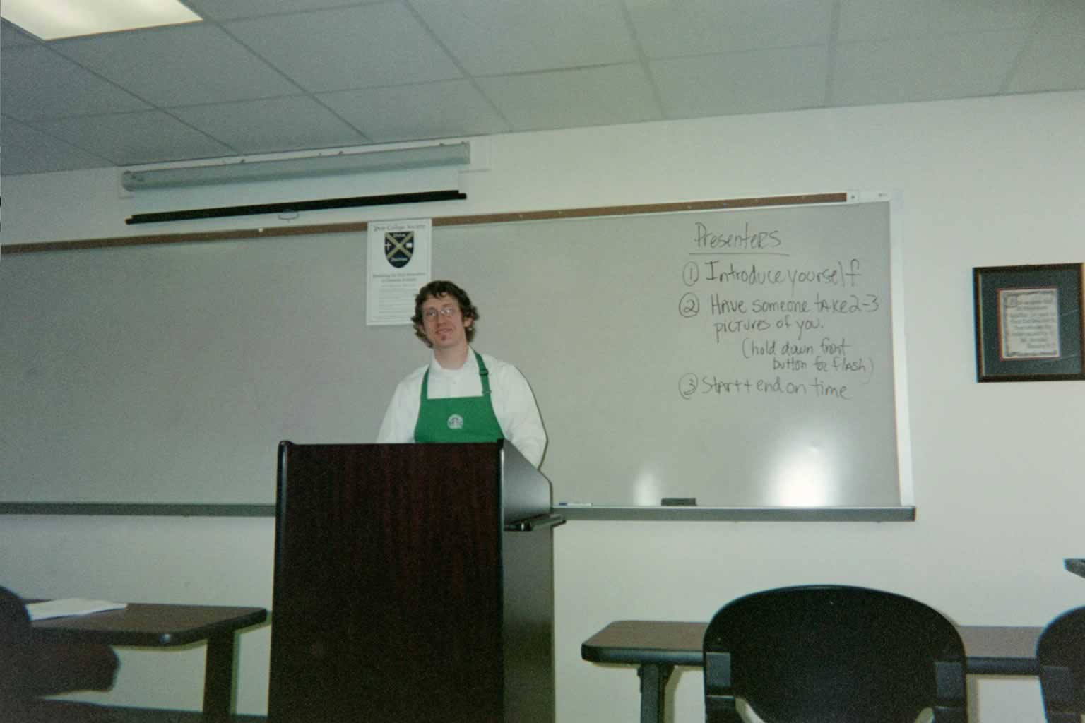 picture of man with glasses standing behind a podium while wearing a green apron