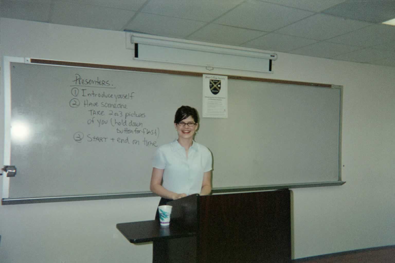 picture of a woman with glasses smiling behind a podium in a classroom