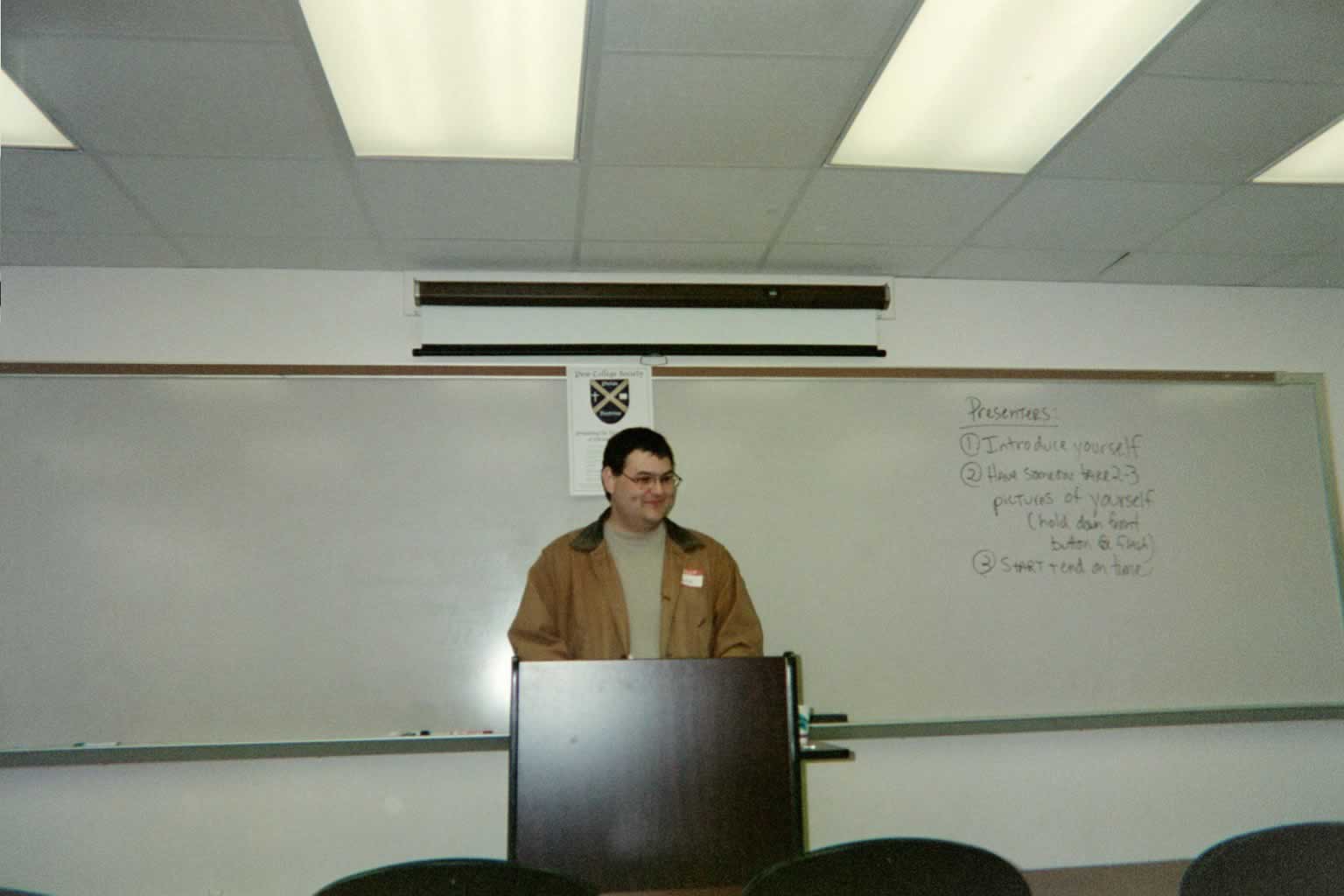 picture of a man in glasses smiling behind a podium in a classroom