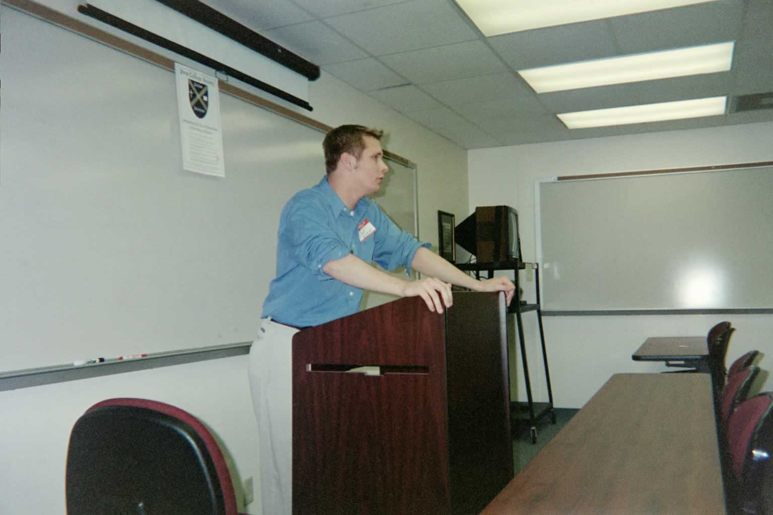 picture of a man in a blue shirt leaning against the podium while speaking