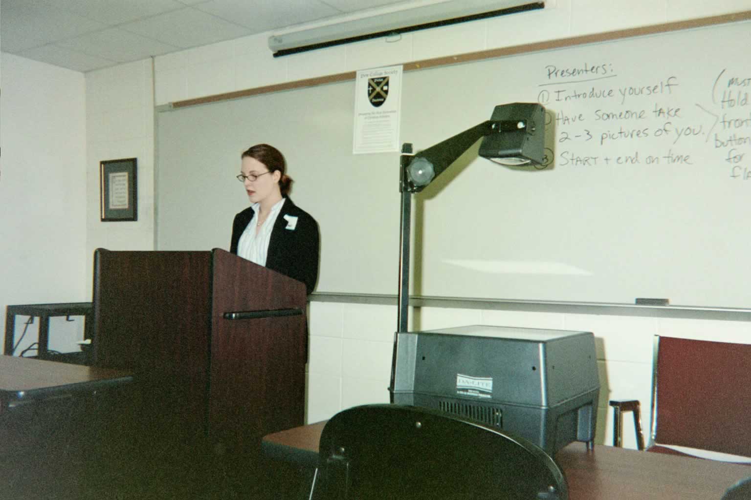 picture of a woman in glasses speaking behind a podium in a classroom