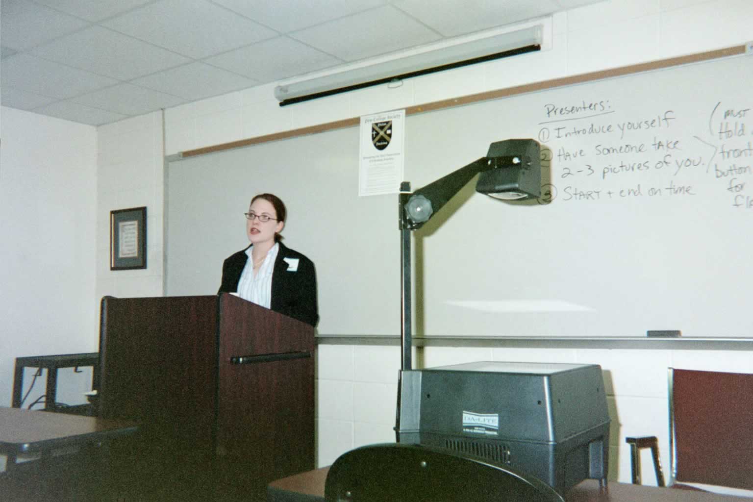 picture of a woman looking up while talking behind a podium in a classroom