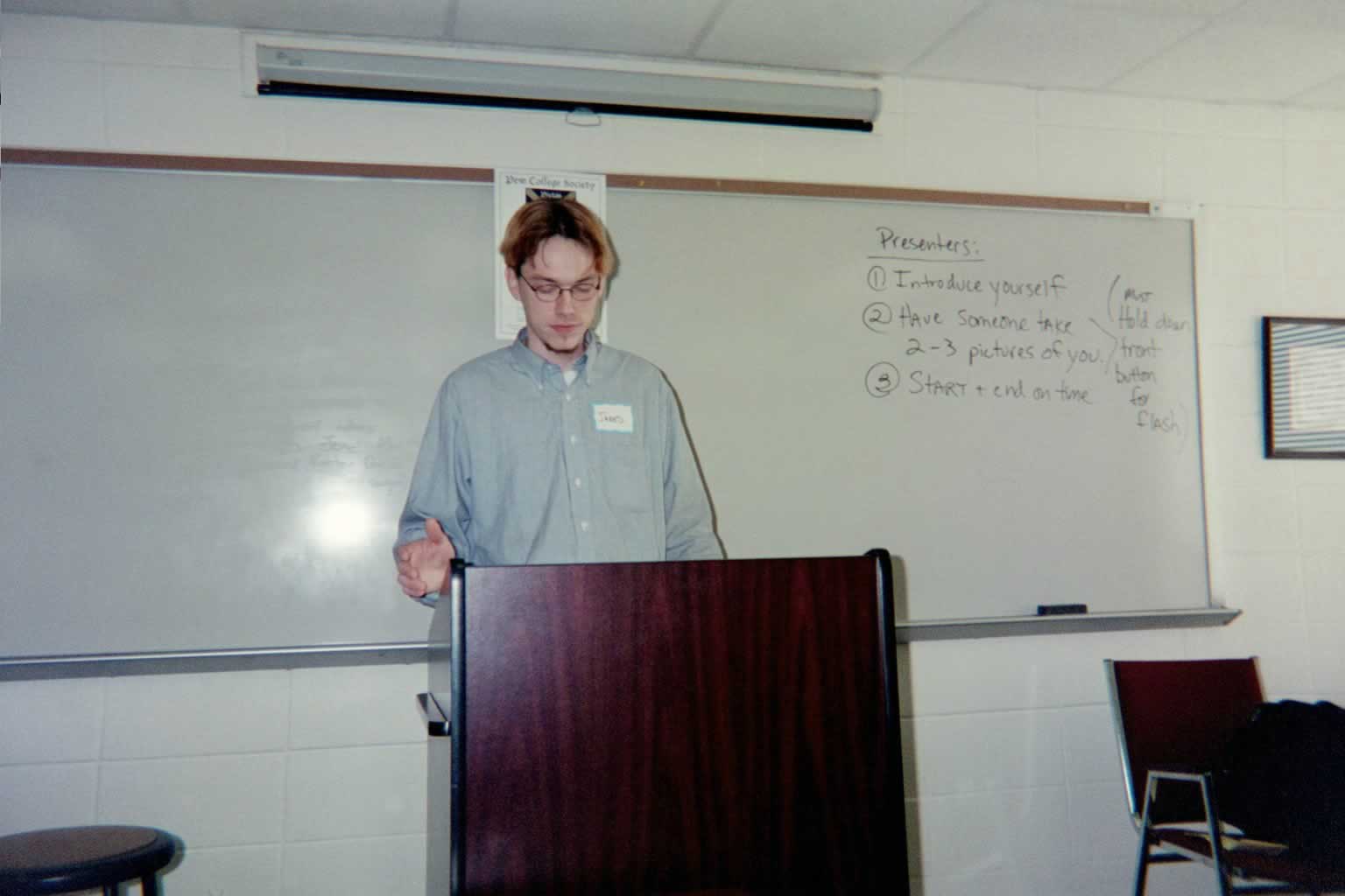 picture of a man wearing a blue shirt with glasses standing behind the podium speaking