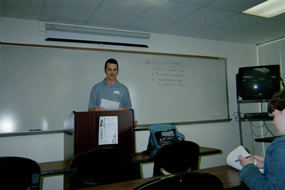picture of a man in a blue shirt standing behind a podium in a classroom speaking
