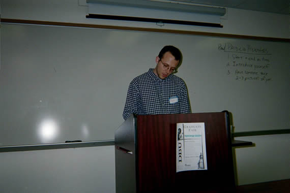 picture of a man in glasses with a checkered shirt standing behind a podium speaking