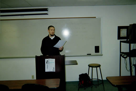 picture of a man standing in the front of a classroom behind a podium holding a piece of paper